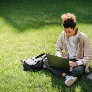 A young man sits on the grass studying with a laptop in a park setting.