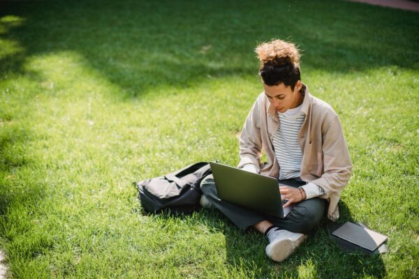 pexels-photo-5553045 A young man sits on the grass studying with a laptop in a park setting.