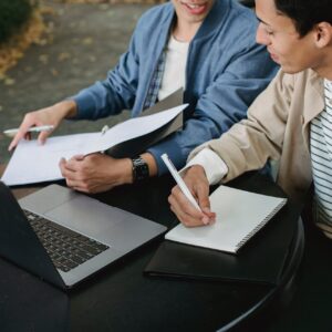 From above of positive male students working on laptop with documents and notebook while doing homework together