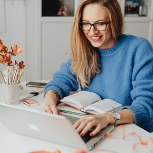 A cheerful woman in a blue sweater working remotely with a laptop in a cozy home setting.